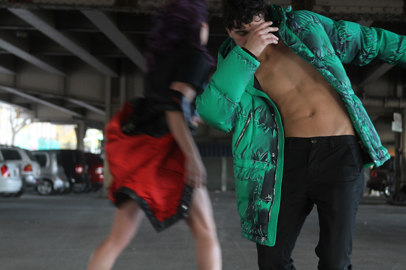 Joey Loto dances in a parking lot underpass. He wears a bright green winter coat and is shirtless underneath. There is another figure in the background and we only see her red skirt.